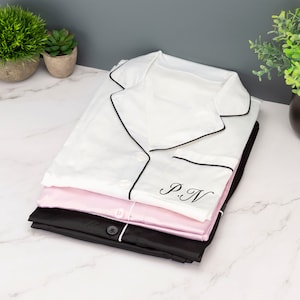 Puede incluir: Una pila de tres pijamas blancos, rosas y negros con ribetes negros y un monograma "P.N." en la parte superior blanca.