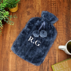 K&ouml;nnte beinhalten: Eine marineblaue Pl&uuml;sch-W&auml;rmflasche mit Bommeln und den gestickten Initialen "R.G." auf der Vorderseite.
