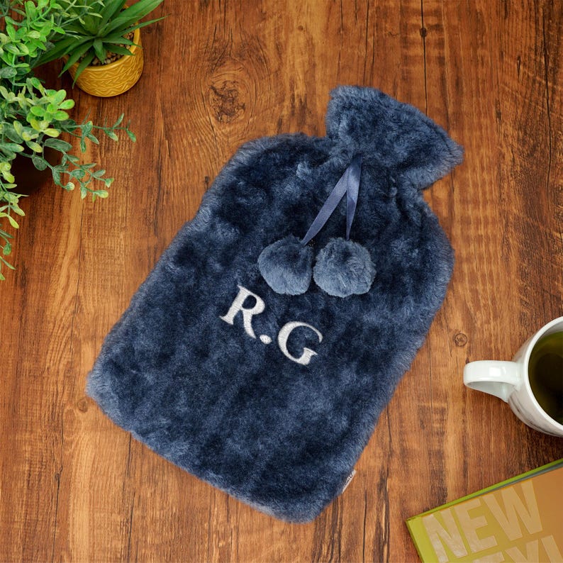 K&ouml;nnte beinhalten: Eine marineblaue Pl&uuml;sch-W&auml;rmflasche mit Bommeln und den gestickten Initialen "R.G." auf der Vorderseite.