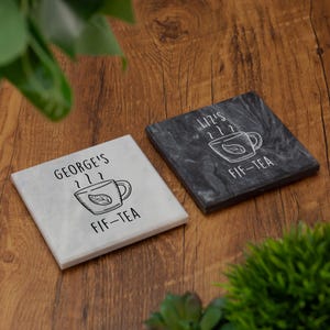 May include: Two square coasters, one white and one dark gray, with a tea cup design. The white coaster says "George's FIF-TEA" and the dark gray coaster says "Liz's FIF-TEA". Both coasters have a tea cup graphic.