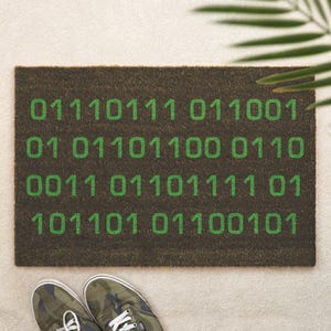 Könnte beinhalten: Eine rechteckige Fußmatte mit dunkelbraunem Hintergrund und grünen Binärcode-Zahlen. Die Fußmatte besteht aus einem groben Material. Tarnfarbene Turnschuhe sind unten im Bild zu sehen.
