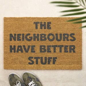 Puede incluir: Felpudo rectangular de fibra de coco marrón natural con la frase "THE NEIGHBOURS HAVE BETTER STUFF" en letras negras. Se ven zapatillas deportivas con estampado de camuflaje en la parte inferior.