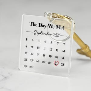 May include: Clear acrylic keychain with a calendar design. The text reads "The Day We Met - September 2021." The date September 27 is highlighted with a red heart. Attached to a gold-colored key ring.