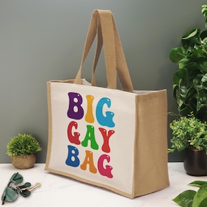 Op de afbeelding: Een rechthoekige draagtas gemaakt van natuurlijke beige jute stof met witte zijpanelen. De voorkant is voorzien van de tekst BIG GAY BAG in grote, bubbelachtige, veelkleurige letters in paars, geel, blauw, rood, groen en roze, met twee stevige handvatten.