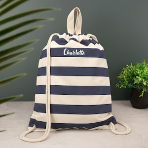 Op de afbeelding: Een rugzak met trekkoord met horizontale strepen in marineblauw en gebroken wit. De tas heeft een crèmekleurig trekkoord en een handvat aan de bovenkant. De naam "Charlotte" staat in witte letters op de voorkant. De tas is van canvas.