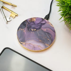 May include: Circular wireless charger featuring a purple and gold abstract pattern. The charger has a wooden edge and a black charging cable. A smartphone, keys, and a small plant are visible.