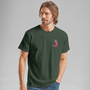 K&ouml;nnte beinhalten: Dunkelgr&uuml;nes T-Shirt mit einer kleinen Grafik auf der linken Brust. Die Grafik ist ein stilisiertes Design in Rot und Grau, das eine geb&auml;ude&auml;hnliche Form und Text darunter zeigt. Das Shirt hat kurze &Auml;rmel und einen Rundhalsausschnitt.