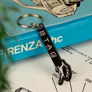 May include: Black keychain with the word "STAG" in white text. The keychain has a ring and a stylized design of a stag's head. The background includes a blue box with the word "FIRENZA" and technical diagrams.