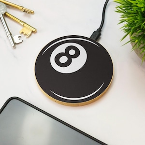 May include: A circular wireless charging pad featuring a black and white graphic of an eight ball. The device has a gold-coloured metallic edge and is connected to a black power cable, designed to charge mobile devices through induction technology.