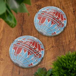 May include: Two round coasters with a globe design. The coasters feature a light blue background with red continents and a silver outline. The word "Trip" is written across the globe in a silver font. The coasters are on a wooden surface.
