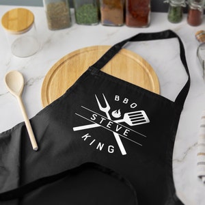May include: Black BBQ apron with white text that reads "BBQ Steve King" and a graphic of crossed grilling tools with a flame.
