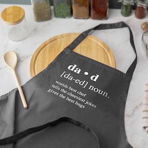 以下が含まれることがあります： 「dad [da-ed] noun. worlds best chef tells the cheesiest jokes gives the best hugs」と白文字で書かれたグレーのキッチンエプロン
