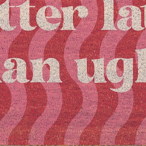 May include: A doormat with a wavy pattern in shades of red and pink. The text "better late than ugly" is displayed in a white, retro font. The mat is made of a textured material.