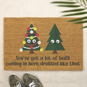 May include: A rectangular doormat made of natural fibres with two cartoon Christmas trees and the text "You've got a lot of balls coming in here dressed like that". The trees have googly eyes and are decorated with baubles.