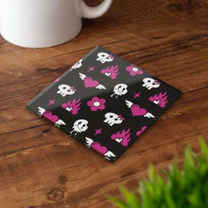 May include: Square black coaster with a repeating pattern of white skulls with pink bows, pink winged hearts, pink flames, and pink flowers. The coaster is on a wooden surface, with a white mug in the background.