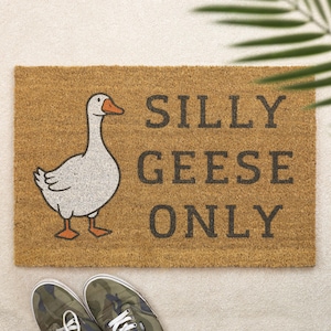 May include: A rectangular doormat made of brown coir fiber. It features a cartoon goose illustration and the text "SILLY GEESE ONLY" in dark gray letters. Camouflage sneakers are visible at the bottom.