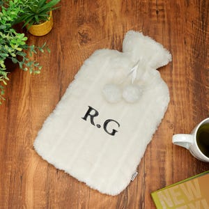 K&ouml;nnte beinhalten: Wei&szlig;e Pl&uuml;sch-W&auml;rmflasche mit einem schwarzen gestickten Monogramm "R.G." und zwei wei&szlig;en Bommeln.