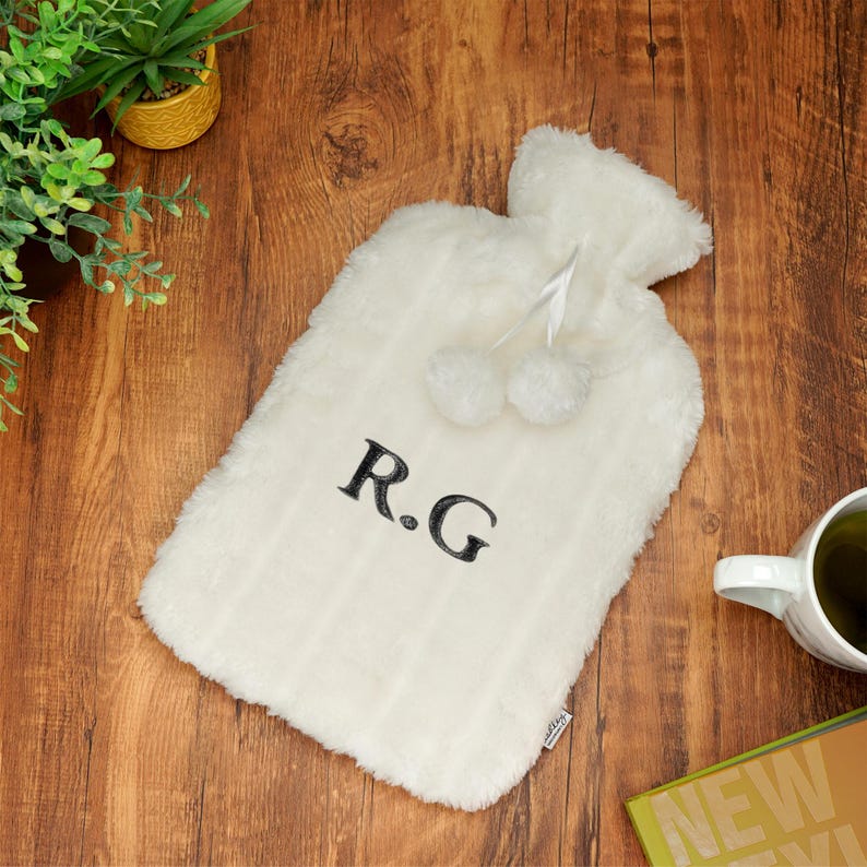 K&ouml;nnte beinhalten: Wei&szlig;e Pl&uuml;sch-W&auml;rmflasche mit einem schwarzen gestickten Monogramm "R.G." und zwei wei&szlig;en Bommeln.