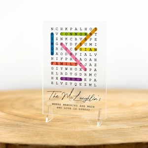 May include: A clear acrylic sign with a colorful word search puzzle featuring the words "The Mclaughlin's", "Family", "Connor", "Simon", "Gemma", and "Prince". The sign also includes the text "Where memories are made and love is spread".