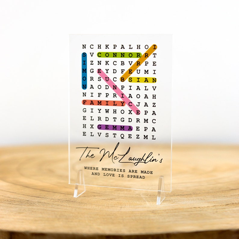 May include: A clear acrylic sign with a colorful word search puzzle featuring the words "The Mclaughlin's", "Family", "Connor", "Simon", "Gemma", and "Prince". The sign also includes the text "Where memories are made and love is spread".