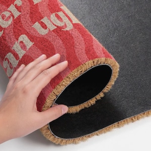 May include: Rolled-up red and beige doormat with the words "You Can't Sit With Us" in beige lettering. The mat has a wavy striped pattern and a fringed edge. The backing is black.