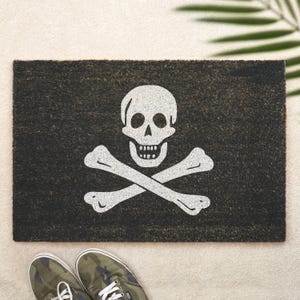 Peut inclure: Tapis de porte rectangulaire noir avec un motif de tête de mort et d'os croisés blancs. Le tapis est fait d'un matériau foncé et texturé. Des baskets de camouflage sont visibles en bas de l'image.