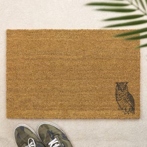 May include: A rectangular doormat made of natural brown coir fiber. A detailed black illustration of an owl is on the lower right corner. The mat is placed on a light beige surface, with a pair of camouflage sneakers visible at the bottom.