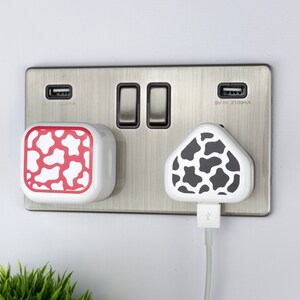 Op de afbeelding: Twee witte stopcontact-adapters met verschillende dierenprintontwerpen. Een adapter heeft een rood-witte koe-printontwerp. De andere adapter heeft een zwart-witte koe-printontwerp. De adapters zijn in een zilveren stopcontact gestoken.