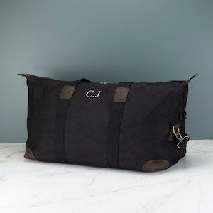 May include: A black canvas duffel bag with brown leather accents and a zip closure. The bag has two top handles and a shoulder strap. The initials "C.J." are embroidered on the front of the bag.