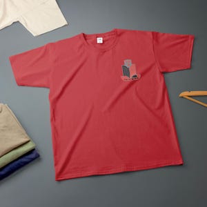 K&ouml;nnte beinhalten: Ein rotes T-Shirt mit einem kleinen Grafikdesign auf der linken Brustseite. Das Design ist stilisiert mit schwarzen, roten und grauen Elementen, und das Wort "TRIUMPH" ist darunter gedruckt. Andere gefaltete Hemden sind sichtbar.