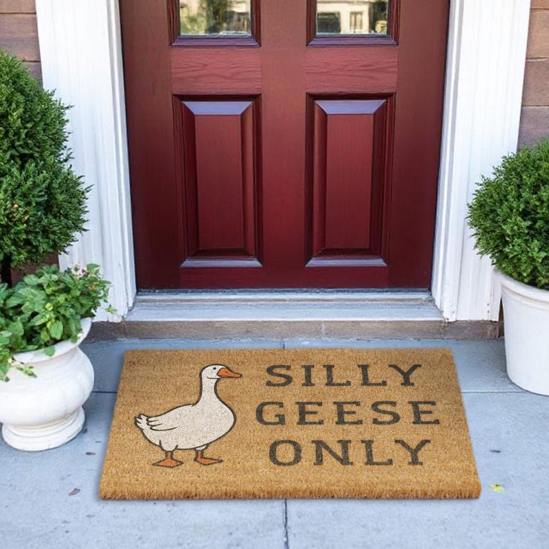May include: A rectangular doormat made of natural fibers, featuring a cartoon goose illustration and the text "SILLY GEESE ONLY" in dark gray. The mat is placed in front of a burgundy door.