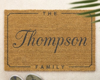 Puede incluir: Una alfombra rectangular de fibra de coco natural. La alfombra presenta el apellido "Thompson" en una fuente elegante, con "THE" arriba y "FAMILY" abajo, todo en negro. Un borde negro enmarca el texto.