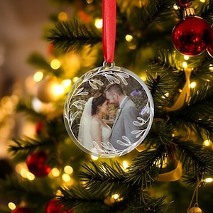 Può includere: Un addobbo natalizio rotondo trasparente con un motivo floreale bianco e una foto di una coppia in abiti da matrimonio all'interno. L'addobbo è appeso a un nastro rosso su un albero di Natale.