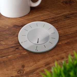 May include: A circular, silver-coloured coaster featuring a dial design. The coaster has a central circular area with a vertical line and surrounding radial lines. The numbers 0 and 11 are visible. A white mug is in the background.