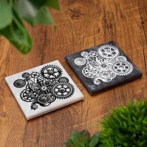 May include: Two square coasters with a steampunk gear design. One coaster is white with a black gear design, and the other is dark gray with a white gear design. The coasters are on a wooden surface.