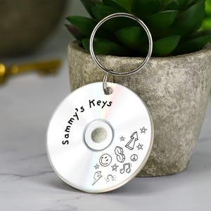 May include: A silver CD-shaped keyring with a metal ring. The keyring features the words "Sammy's Keys" and doodle-style graphics, including a smiley face, musical notes, and an arrow.