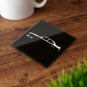 Puede incluir: Posavasos cuadrado negro con un contorno blanco de un coche y el texto "MG ZT". El posavasos está sobre una superficie de madera, con una taza blanca en el fondo.