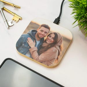 Può includere: Un caricabatterie wireless quadrato con cornice in legno. Il caricabatterie presenta una fotografia a colori di una coppia sorridente. Un cavo di ricarica nero è collegato. Sono visibili anche chiavi e una piccola pianta.