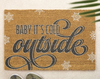 May include: A rectangular doormat with the phrase "BABY IT'S COLD outside" in black script and block letters, with white snowflake accents. The mat is brown and has a textured surface. Camouflage sneakers are at the bottom.