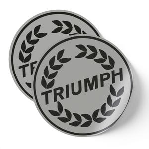 May include: Two silver-colored, circular stickers. Each sticker features the word "TRIUMPH" in black, encircled by a black laurel wreath. The stickers are slightly overlapping, with a white background.