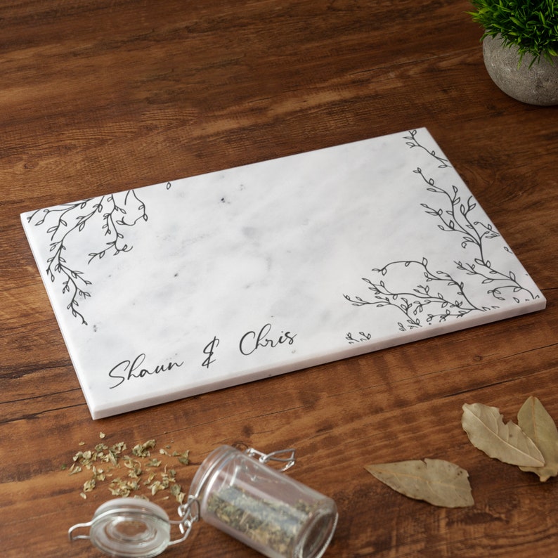Puede incluir: Tabla de servir de m&aacute;rmol blanco con dise&ntilde;o floral negro y los nombres "Shaun & Chris" escritos en cursiva.