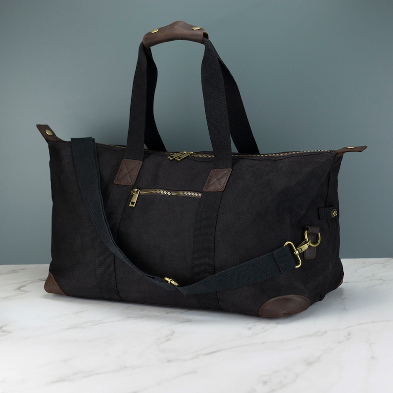 May include: A black canvas duffel bag with brown leather accents and a gold zipper. The bag has a long adjustable shoulder strap and two top handles.