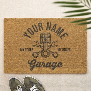 May include: A rectangular doormat made of natural brown coir fibre. The mat features the text "YOUR NAME" arched above a graphic of a piston and spanners. Below the graphic are the words "MY TOOLS" and "MY RULES", and the word "Garage".