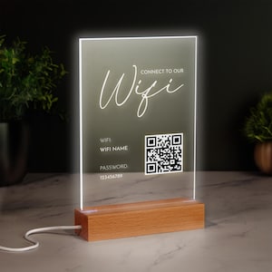 Può includere: Un cartello in acrilico bianco con una base in legno che dice "Connect to our Wifi" con un carattere stilizzato. Il cartello include anche un codice QR e il testo "WIFI: WIFI NAME PASSWORD: 123456789".