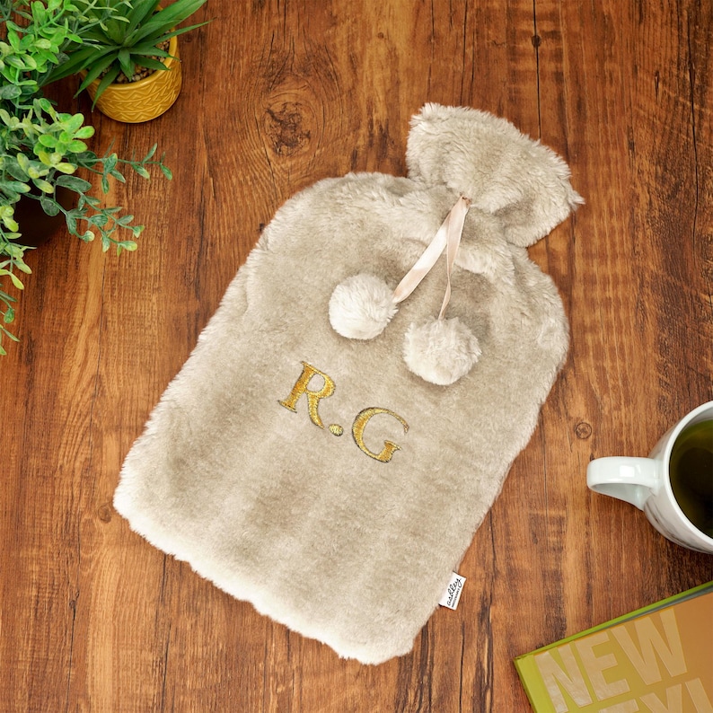 K&ouml;nnte beinhalten: Eine beige Kunstfell-W&auml;rmflasche mit einem cremefarbenen Band und zwei Bommeln. Die Initialen "R.G." sind in Gold auf der Vorderseite gestickt.