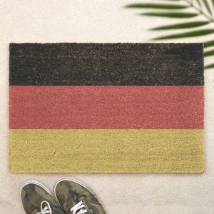 May include: A rectangular doormat featuring the German flag design, with horizontal stripes in black, red, and gold. The mat is made of a textured material. Camouflage sneakers are visible at the bottom.