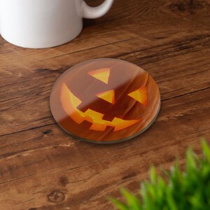 May include: A circular glass coaster with a carved pumpkin design. The pumpkin is orange with a glowing yellow interior, set against a dark brown background. A white mug is in the background.