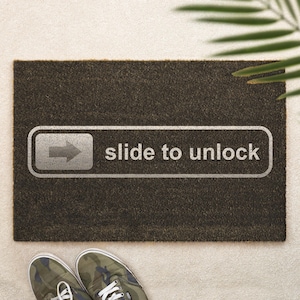 Könnte beinhalten: Eine rechteckige Fußmatte mit einer dunkelbraunen, strukturierten Oberfläche. Die Matte zeigt eine weiße Grafik einer Schiebe-Pfeil-Taste mit den Worten "slide to unlock" in Weiß. Ein Paar Camouflage-Sneaker sind unten.