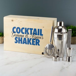May include: A wooden box with the text "Cocktail Gemma & Ryan's Shaker" in blue lettering. The box contains a silver cocktail shaker, a silver cocktail strainer, a silver jigger, a silver bar spoon, and a silver muddler.