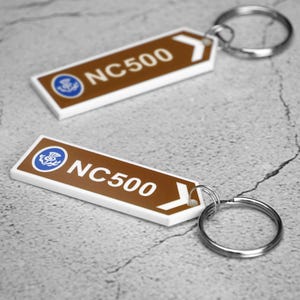 May include: Two rectangular keychains with a brown background and white border. The keychains have the text "NC500" in white, a blue circle with a thistle design, and a white arrow. Each keychain has a silver key ring.